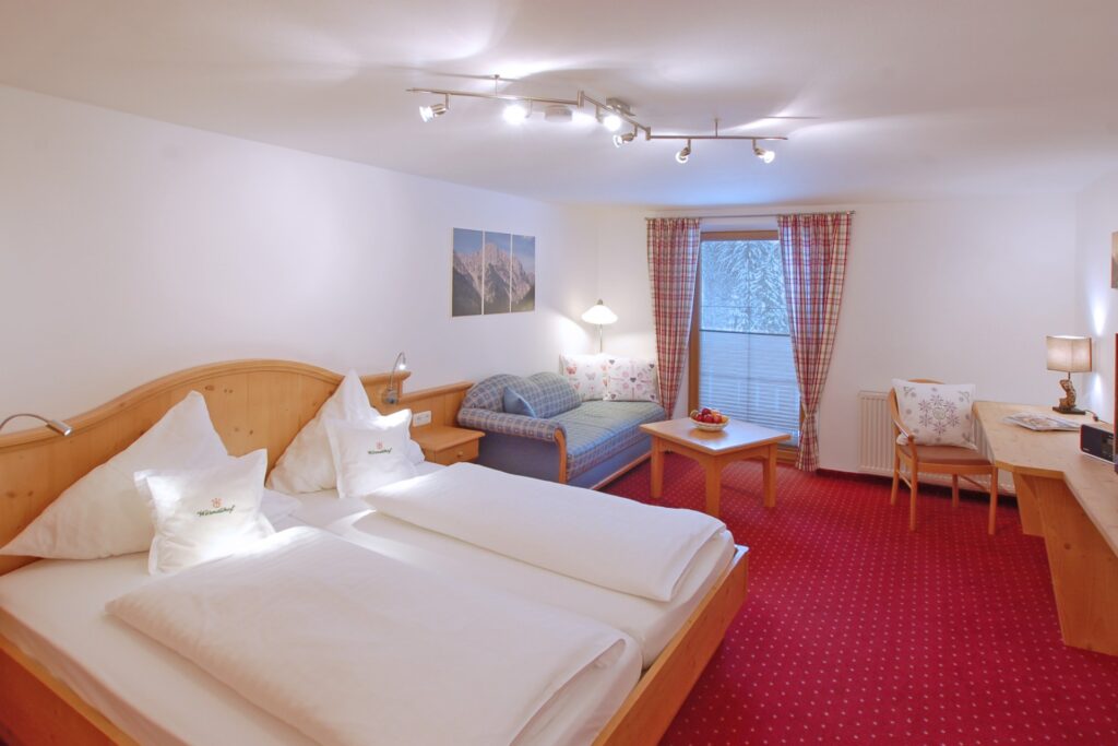 Helles Doppelzimmer im alpinen Stil mit Holzbetten, rotem Teppich, Schreibtisch, Sofa und Zugang zum Balkon im Hotel Wörndlhof Ramsau.