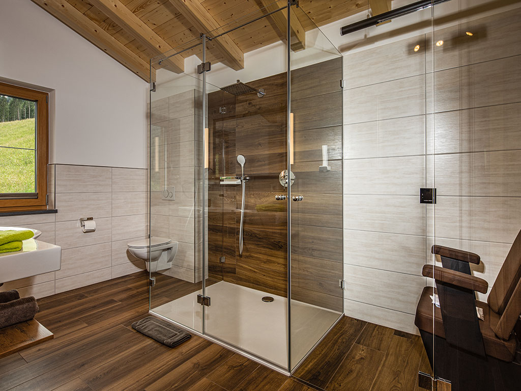 Modernes Badezimmer im Hotel Wörndlhof Ramsau mit bodengleicher Glasdusche, Holzoptik, WC, Waschtisch und Fenster mit Blick ins Grüne.